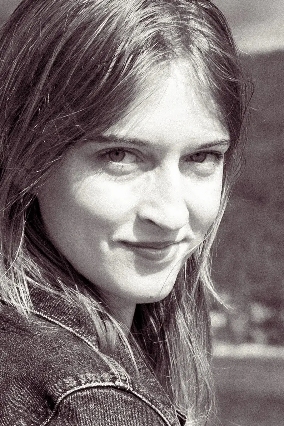 Black-and-white close-up portrait of a woman in a denim jacket, glancing over her shoulder with a soft smile