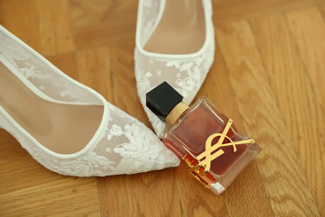 The bride's lace wedding shoes resting on a wood floor beside a bottle of perfume.