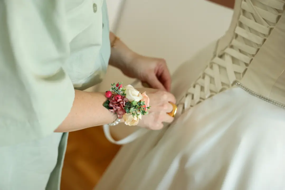 A helper's hands lacing up the back of the wedding gown, a floral wrist corsage in view.