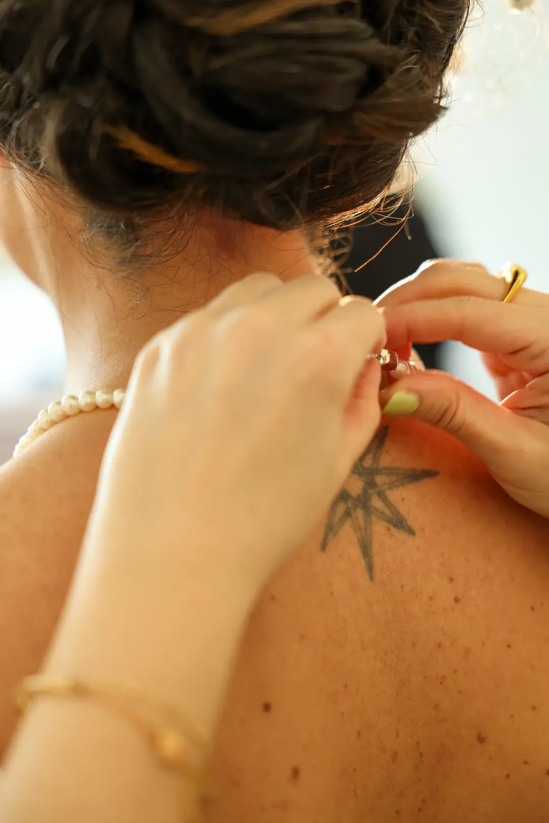 Hands fastening a necklace clasp at the back of the bride's neck, a small star tattoo on her shoulder.