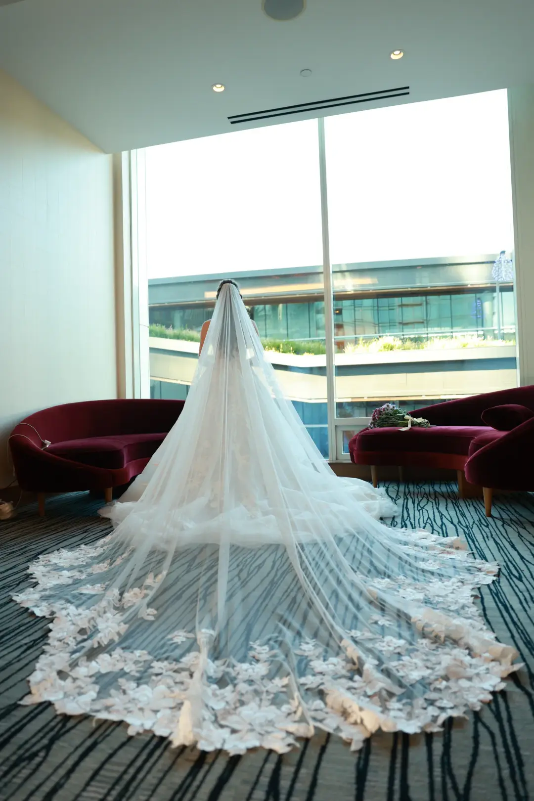 The bride seen from behind in a long cathedral veil, gazing out a window at the city skyline.