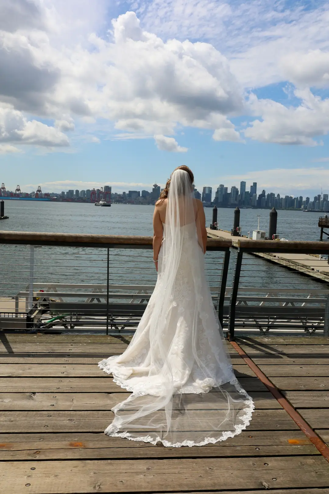 The bride standing on a waterfront pier, her lace train flowing behind her as she views the city skyline.