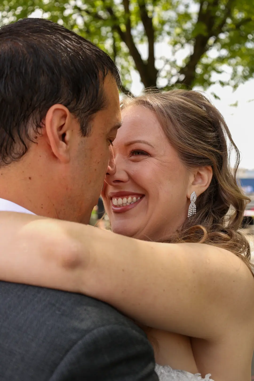 A close, joyful embrace between the newlyweds outdoors beneath green trees.