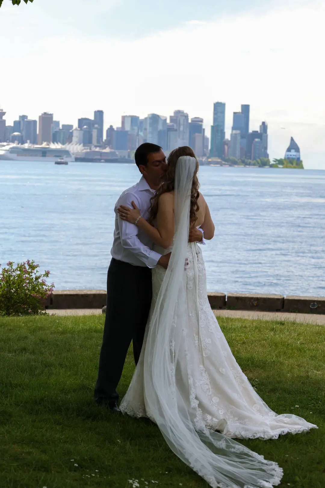 The newlyweds kissing on a waterfront lawn with the city skyline across the water.