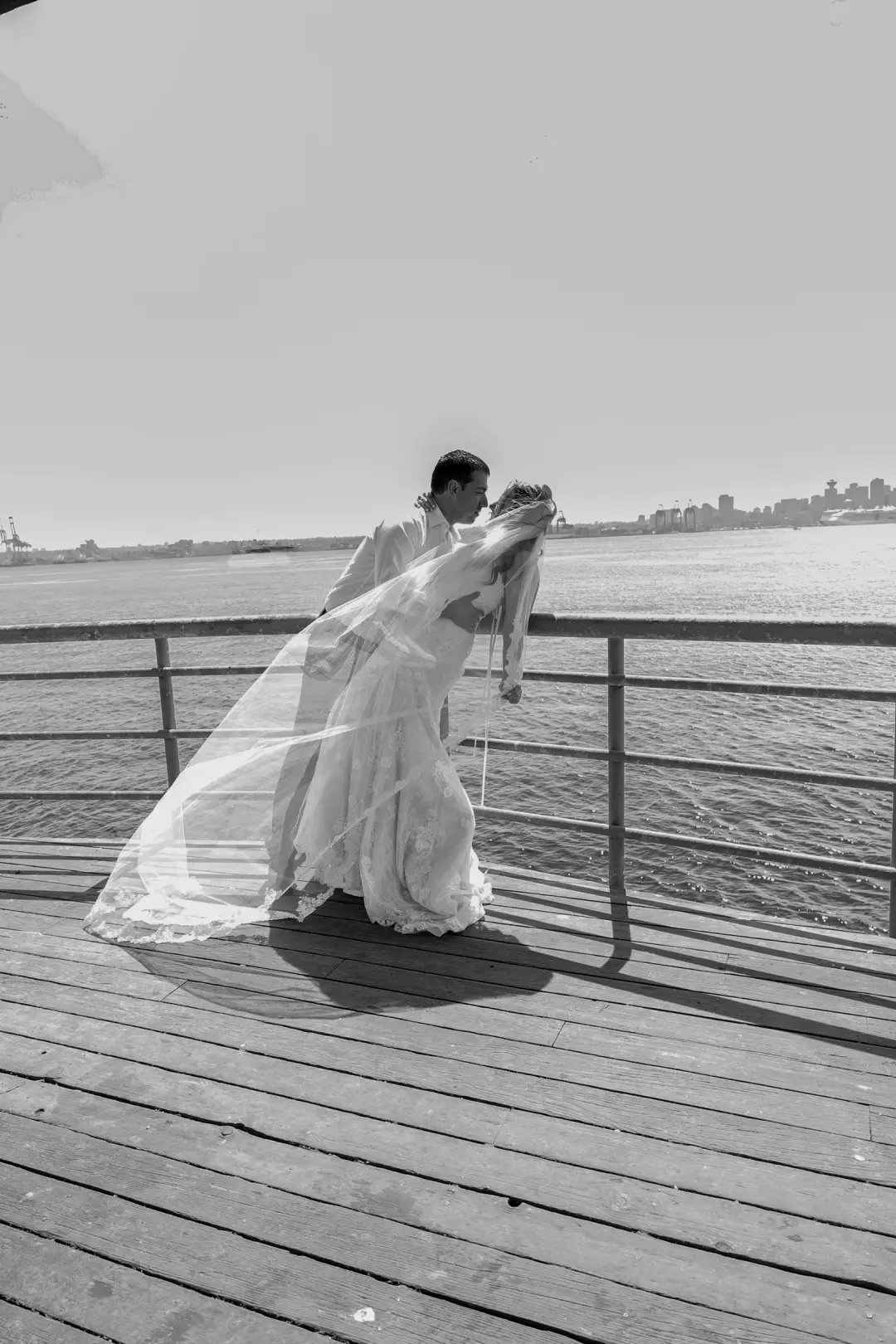 A black-and-white photo of the couple embracing on a pier as the bride's veil billows in the wind.